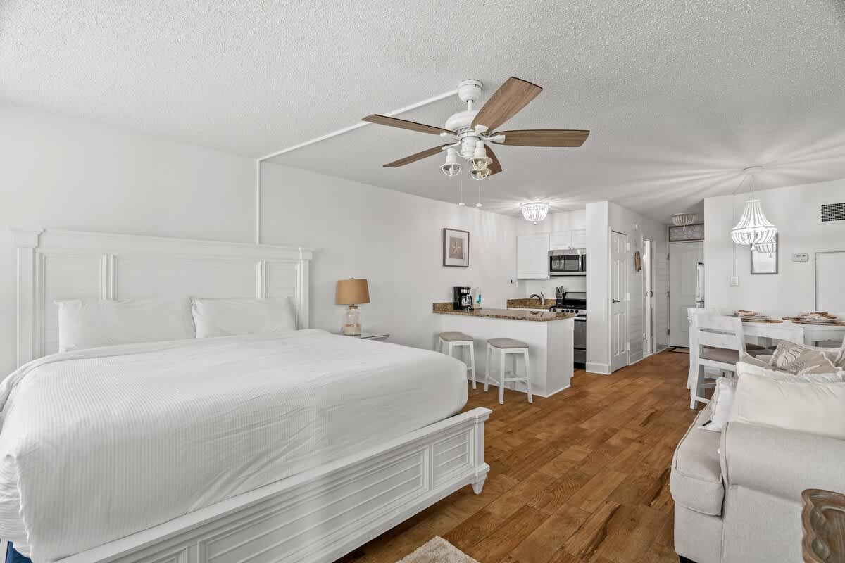 Top Of The Gulf 213 – Compact studio with a white bed, kitchenette, dining area, and wooden flooring.