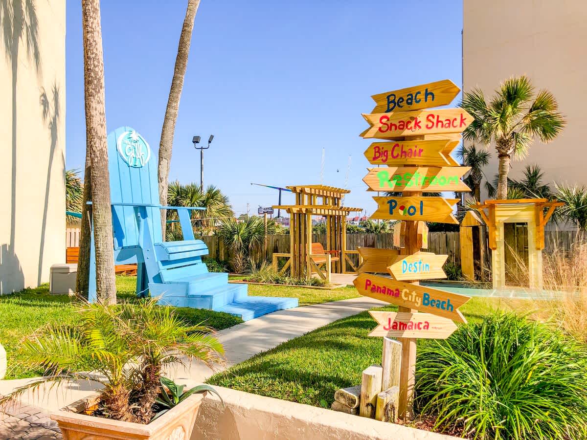 Top Of The Gulf 213 – Blue oversized chair, colorful directional signs, palm trees, and grassy area in a vacation rental outdoor space.