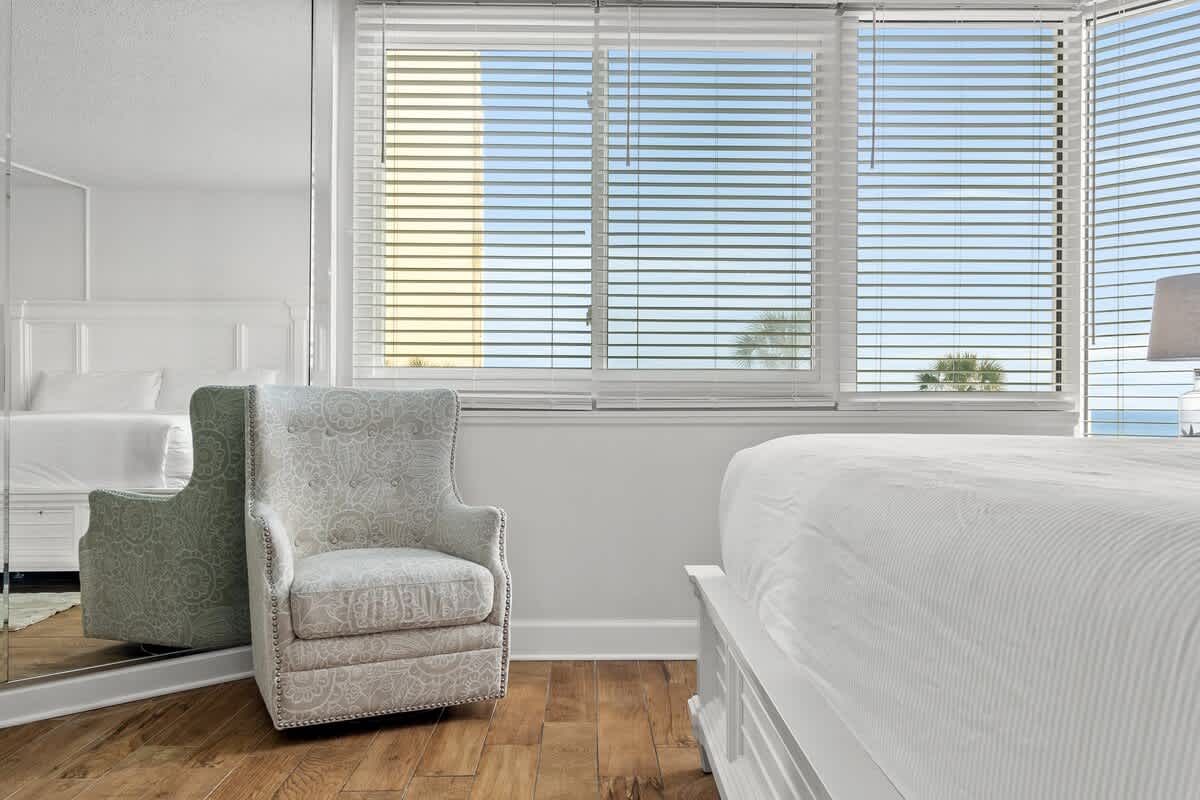 Top Of The Gulf 213 – Bright room with a patterned armchair, bed, and window view of palm trees and blue sky.