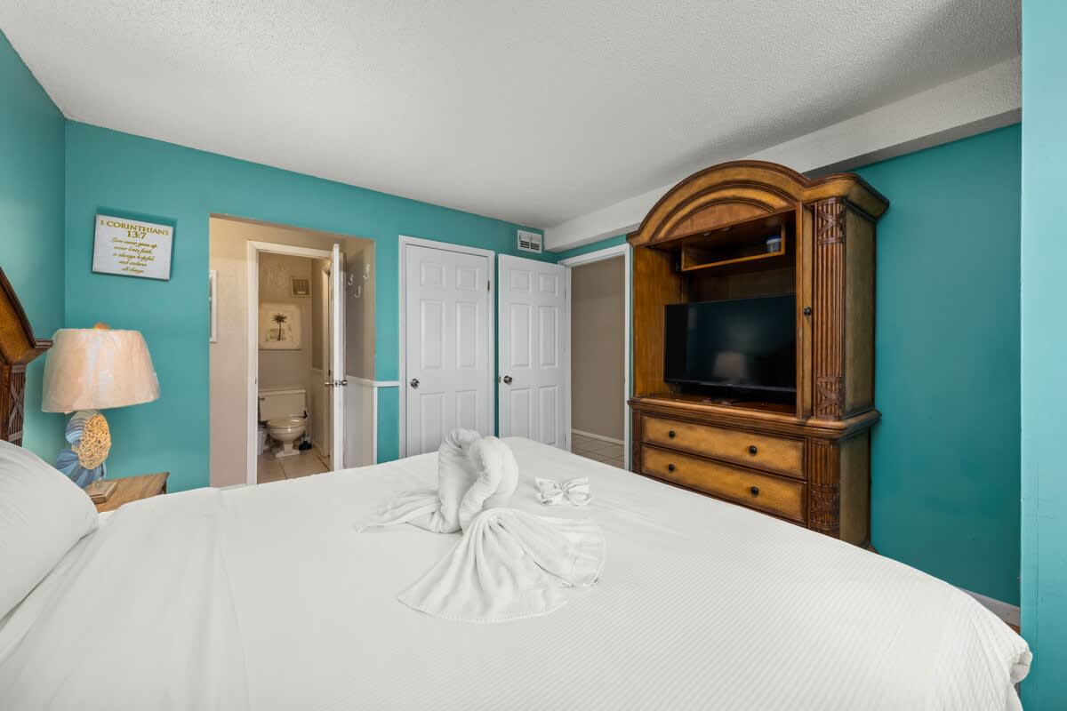 The Summit 1023 – Bedroom with a white bed, a lamp, dresser, and bathroom doorway.