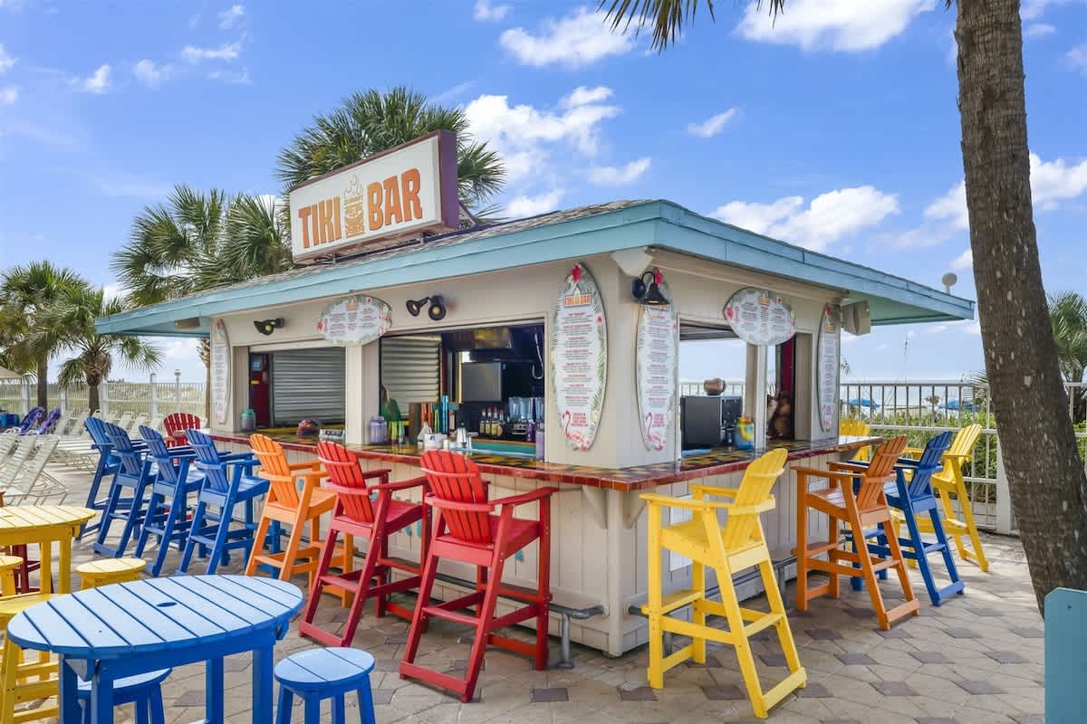 The Summit 616 – Outdoor tiki bar with colorful seating and palm trees, overlooking the beach and blue sky.