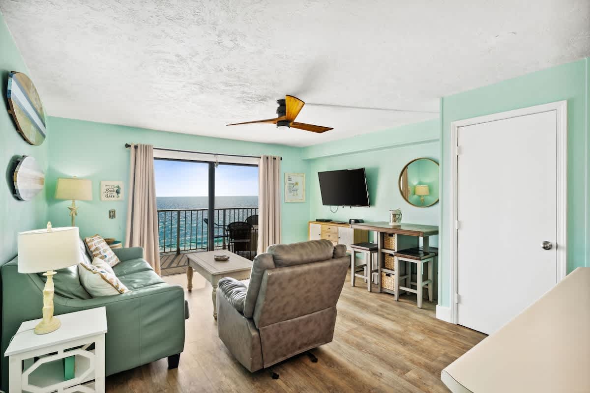 The Summit 1322 – Living room with ocean view, light green walls, sofa, recliner, and small dining area.