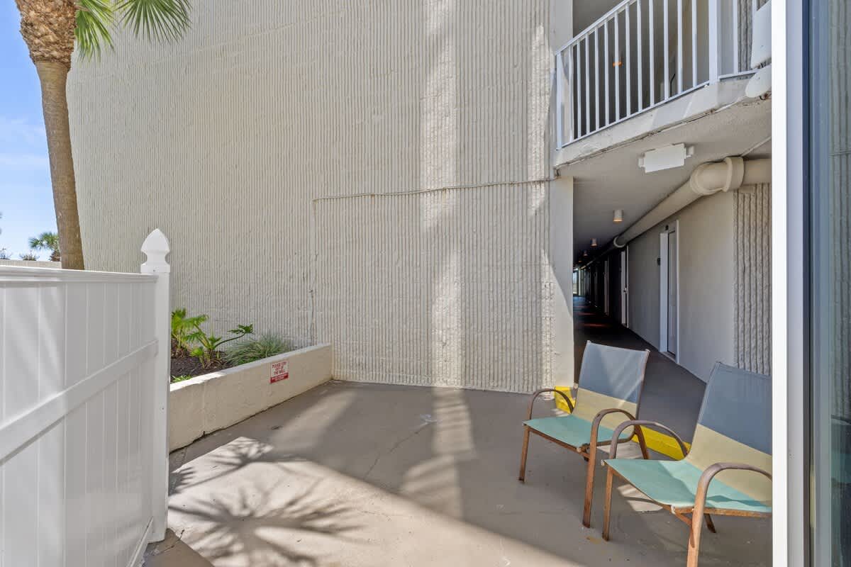 Top Of The Gulf 111 – Two chairs on a patio near a white fence, with a view of a pathway and palm trees.