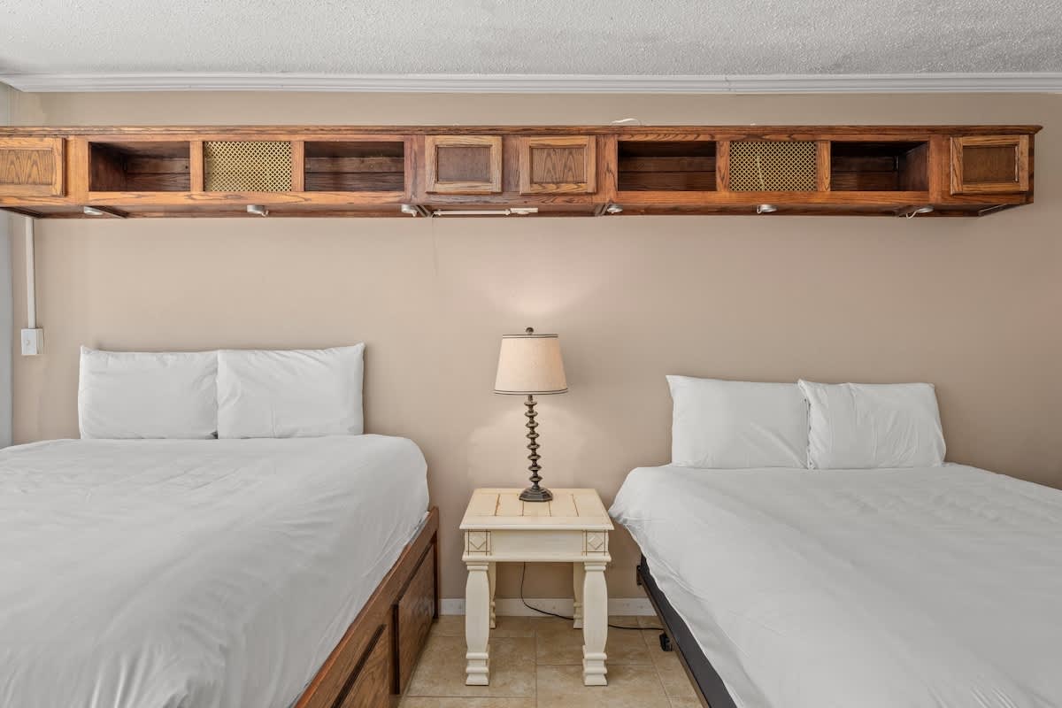 Top Of The Gulf 111 – Two beds with white linens, a bedside table lamp, and overhead storage in a beige room.