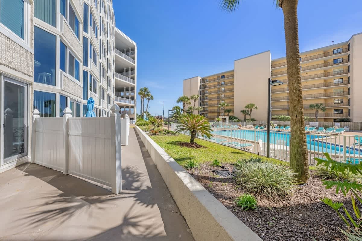 Top Of The Gulf 111 – Path leading to pool area with palm trees and multi-story buildings in the background.