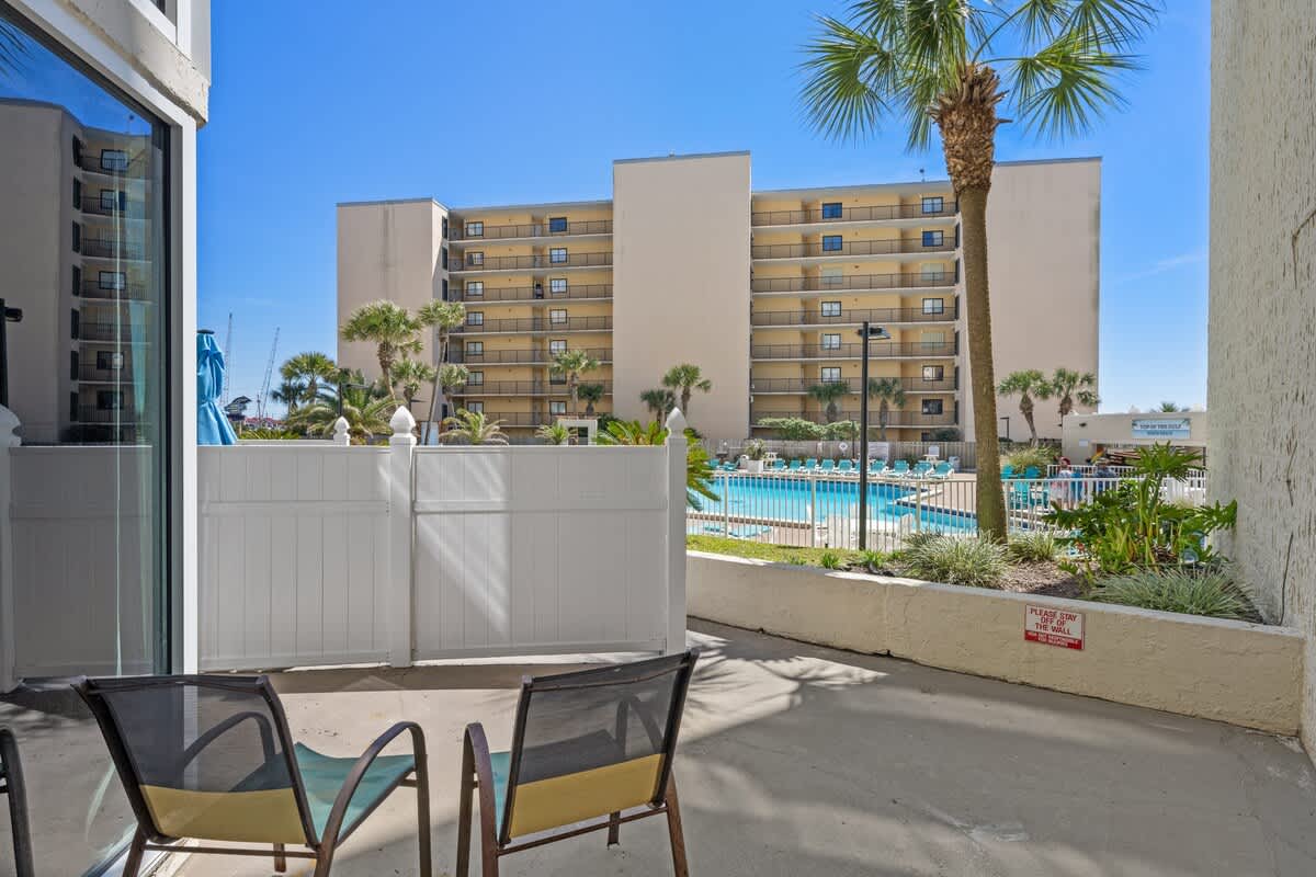 Top Of The Gulf 111 – Patio with two chairs overlooking a pool area and palm trees, adjacent to a multi-story building.