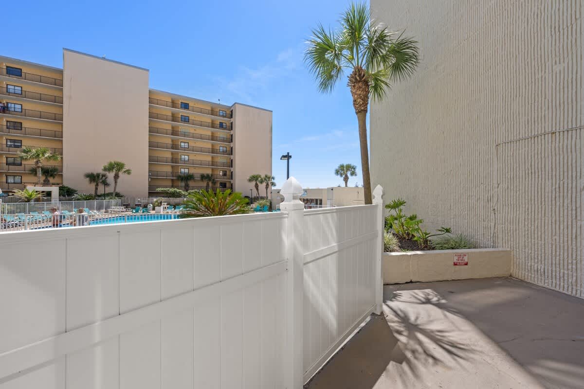 Top Of The Gulf 111 – View of a fenced area with tropical plants and a swimming pool in the background.