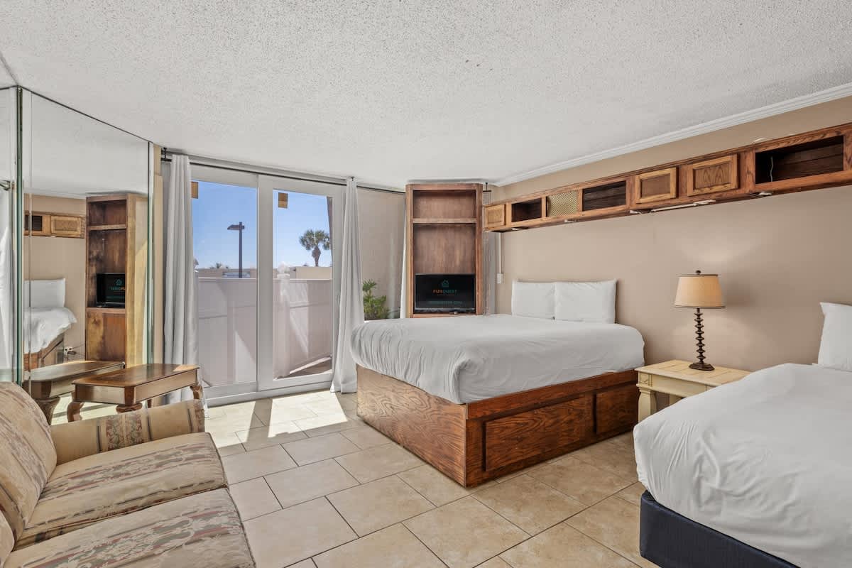 Top Of The Gulf 111 – Cozy vacation rental room with two beds, seating area, large mirrors, and sliding door offering an outdoor view.