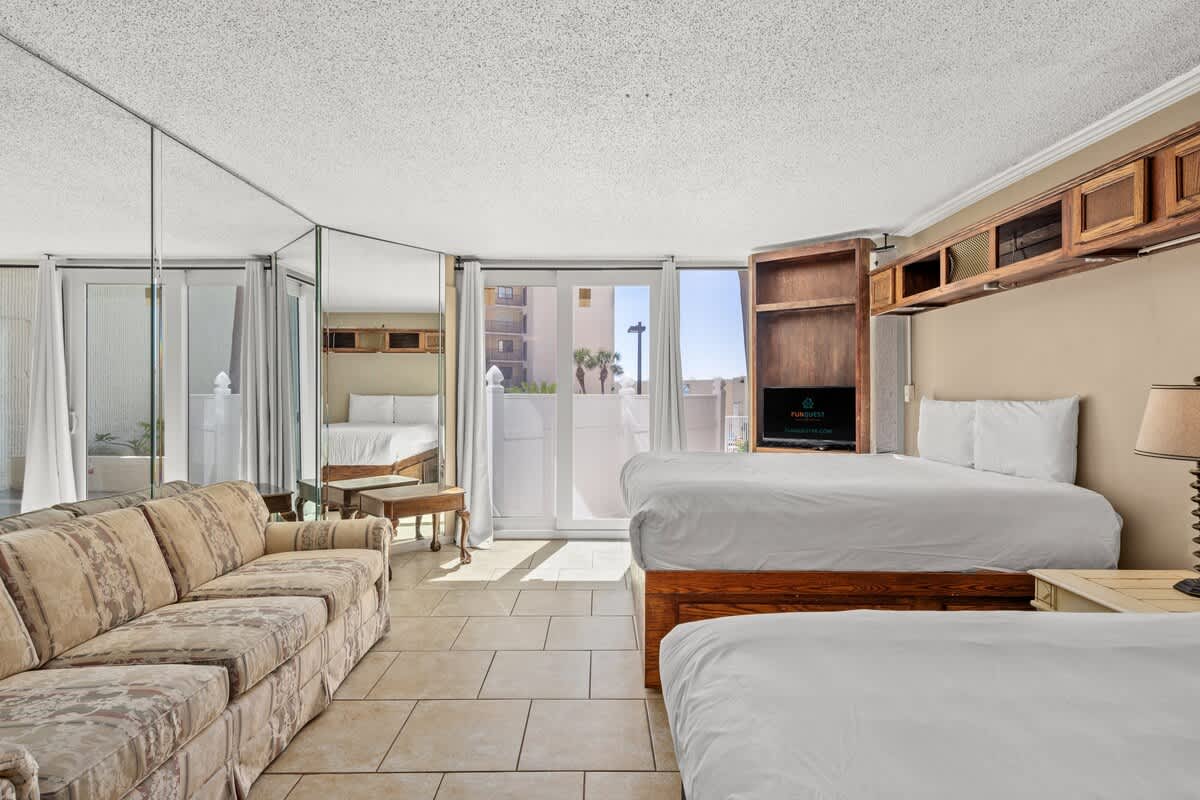 Top Of The Gulf 111 – Studio room with two beds, sofa, and sliding glass doors leading to a small outdoor area.