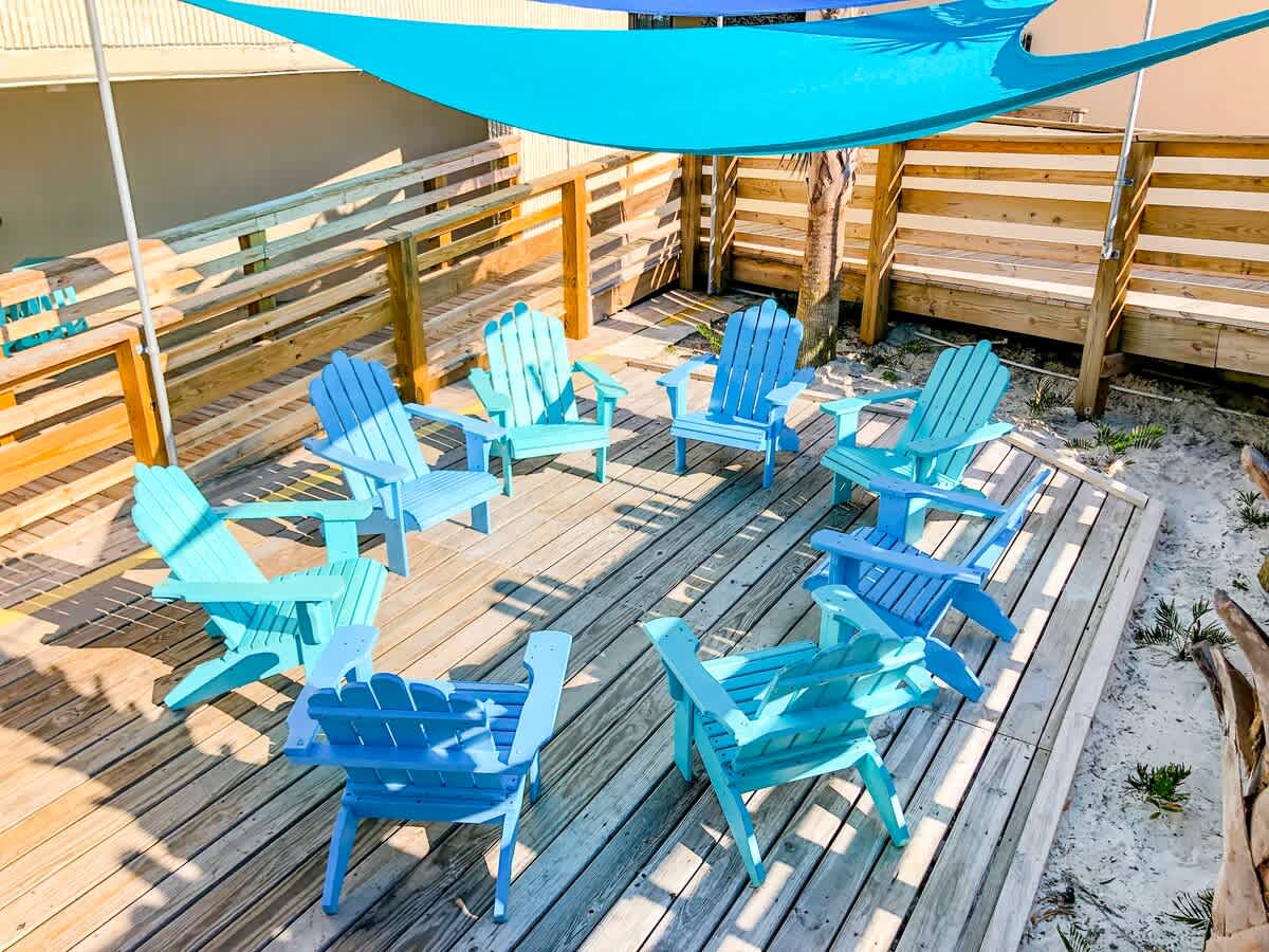 Top Of The Gulf 621 – Outdoor patio area with wooden deck and multiple blue adirondack chairs under a shade sail.