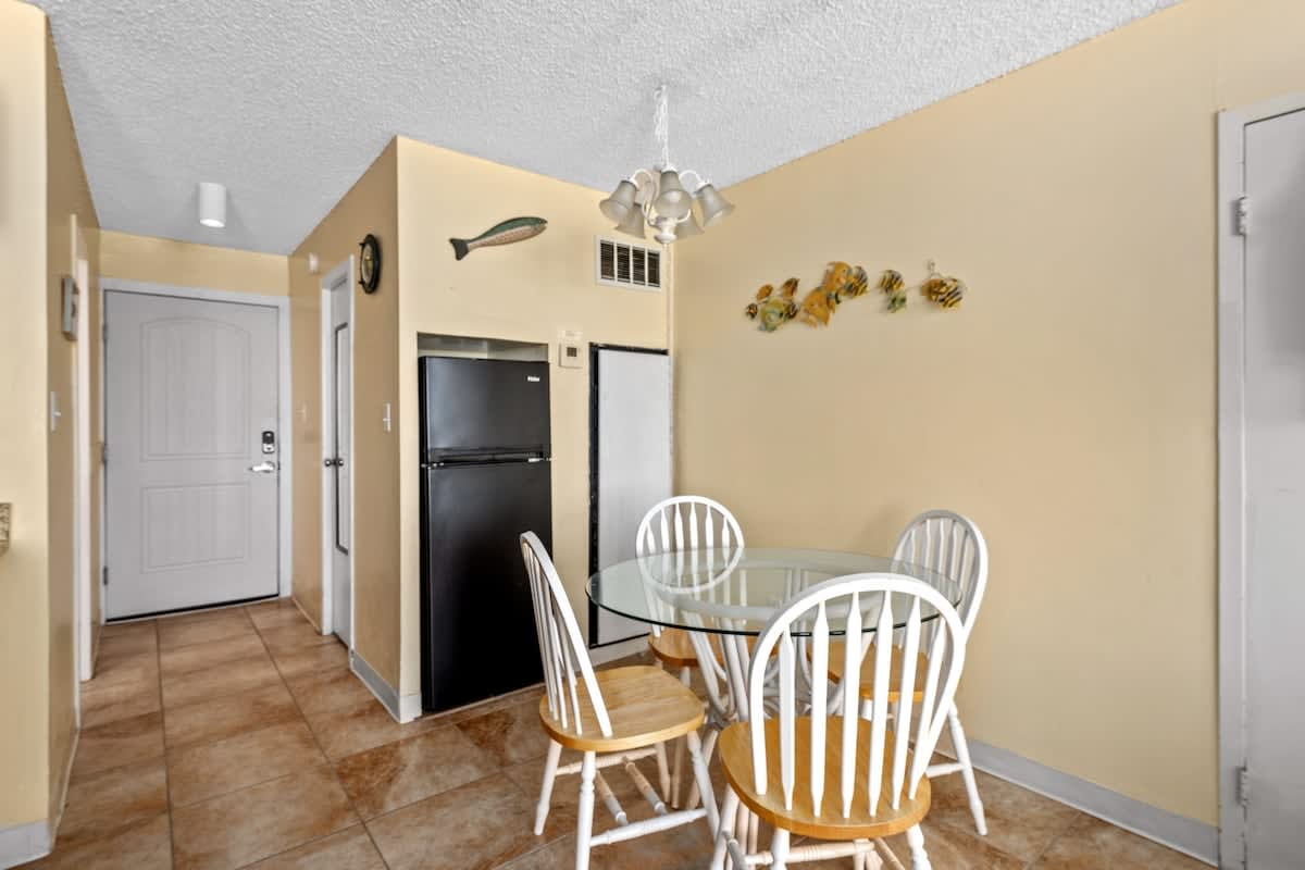 Top Of The Gulf 621 – Dining area with a glass table and four white chairs, featuring a refrigerator and wall decorations.