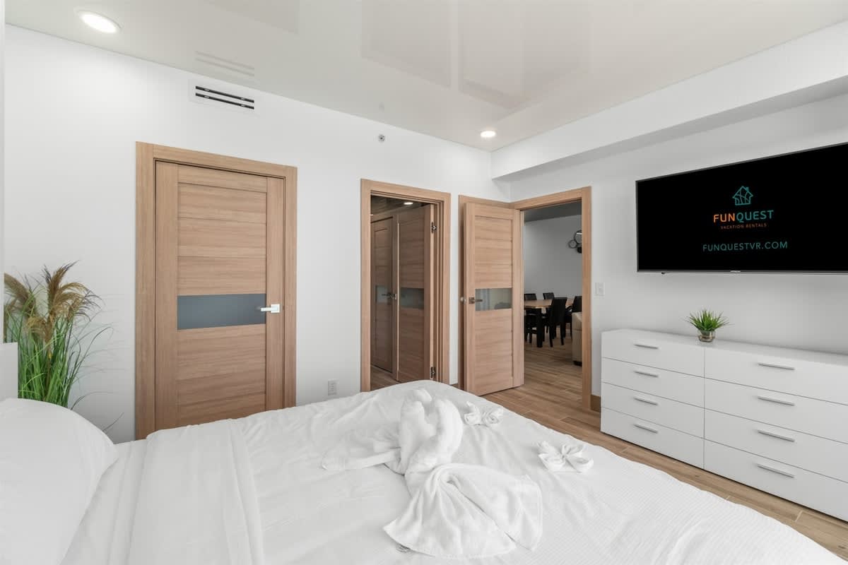 Ocean Villa 1302 – Bedroom with a white bed, wooden doors, a television, and a dining area visible through an open door.