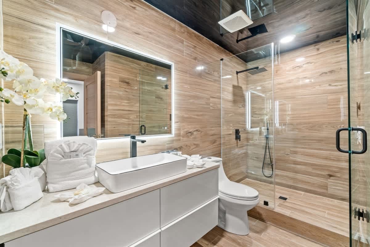 Ocean Villa 1302 – Modern bathroom with wooden accents, glass shower, large mirror, white sink, and decorative plants.