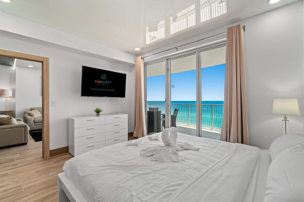 Ocean Villa 1302 – Bedroom with ocean view, large window, white bedding, TV, and seating area visible.