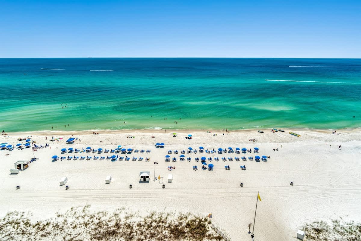 Ocean Villa 1302 – View of the beach with blue umbrellas, lounge chairs, and the turquoise ocean under a clear sky.