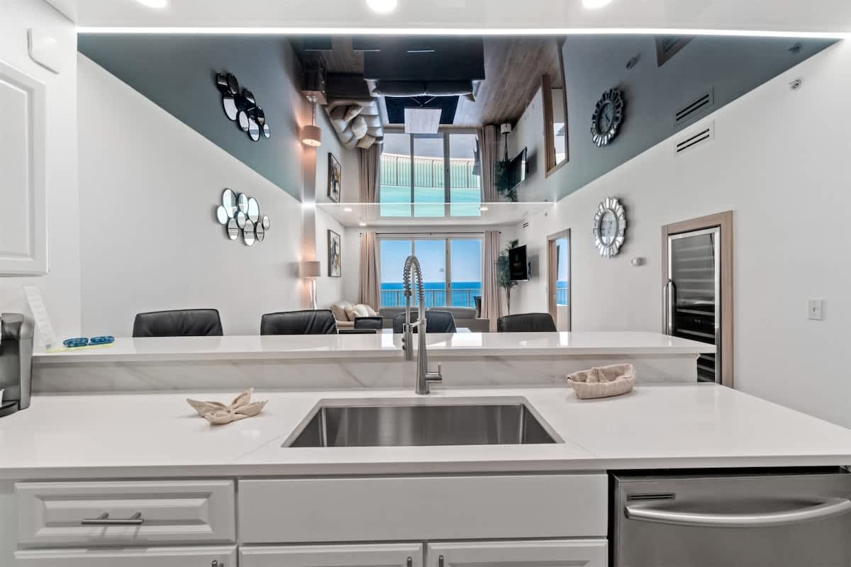 Ocean Villa 1302 – Modern kitchen with white countertops, stainless steel sink, and view of living area and ocean through large windows.