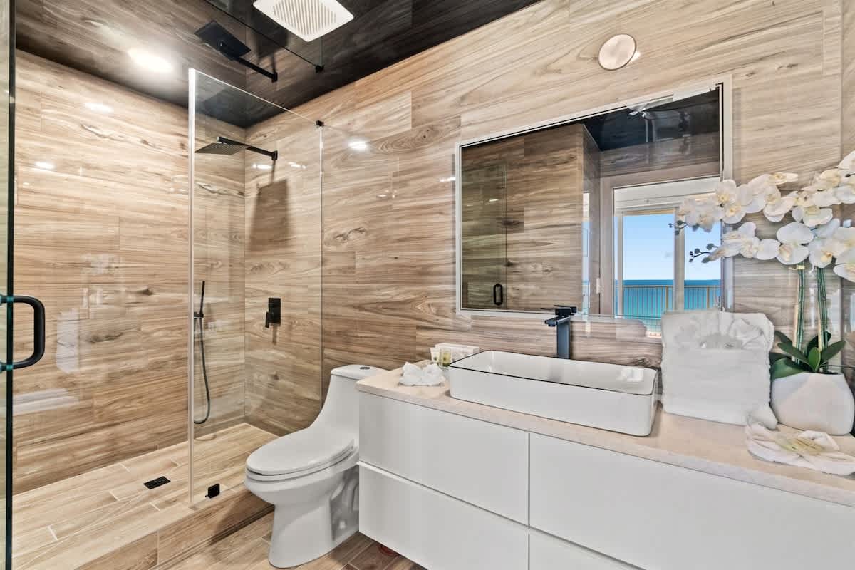 Ocean Villa 1302 – Modern bathroom with a glass shower, white vanity, and ocean view through a window.