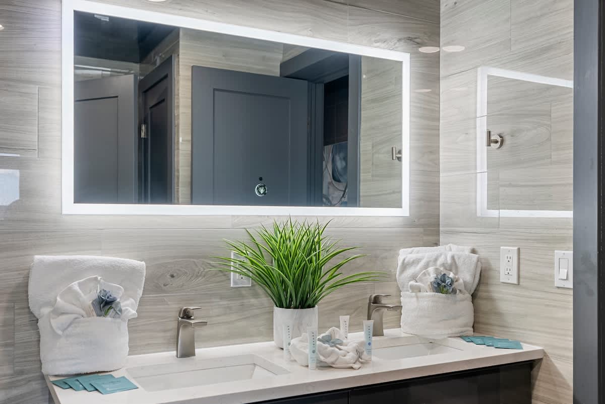 The Summit 1205 – Modern bathroom with double sinks, illuminated mirror, towels, and decorative plant.