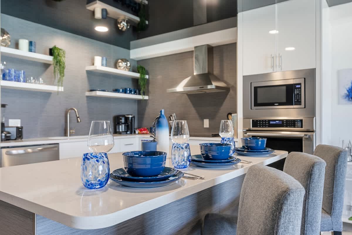 The Summit 1205 – Modern kitchen with quartz countertop, blue dishware, three grey chairs, and stainless steel appliances.