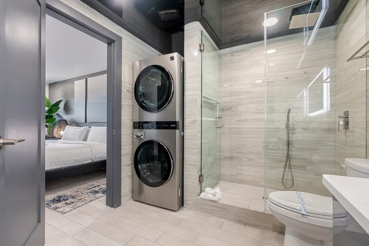 The Summit 1205 – Bathroom with shower, laundry machines, and view into a bedroom with a bed and plant.