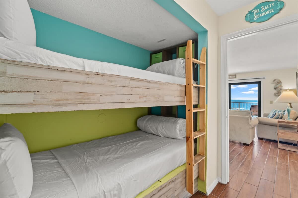 The Summit 731 – Bunk beds with white linens and a turquoise accent wall, view of living area and ocean.