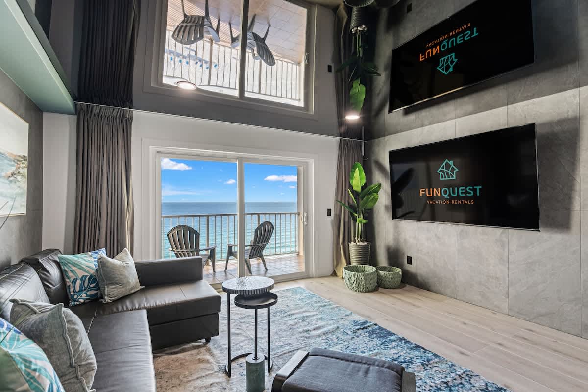 The Summit 1305 – Living room with modern decor, two large TVs, and a balcony view of the ocean.