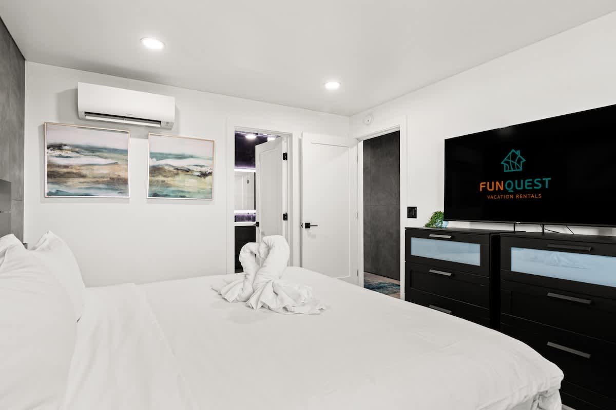 The Summit 1305 – Modern bedroom with a white bed, TV, artwork, and a door leading to another room.