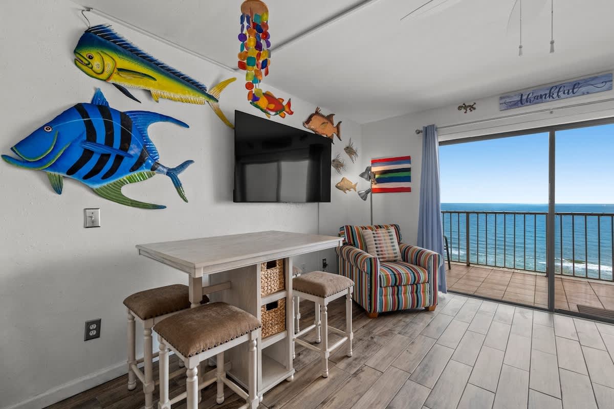 The Summit 1213 – Living area with ocean view, TV, striped chair, dining table, and colorful fish decorations.