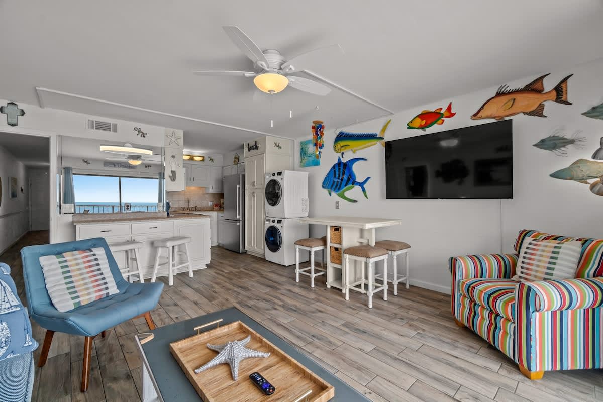 The Summit 1213 – Living room with colorful furniture, a TV, kitchen area, and ocean view through large window.