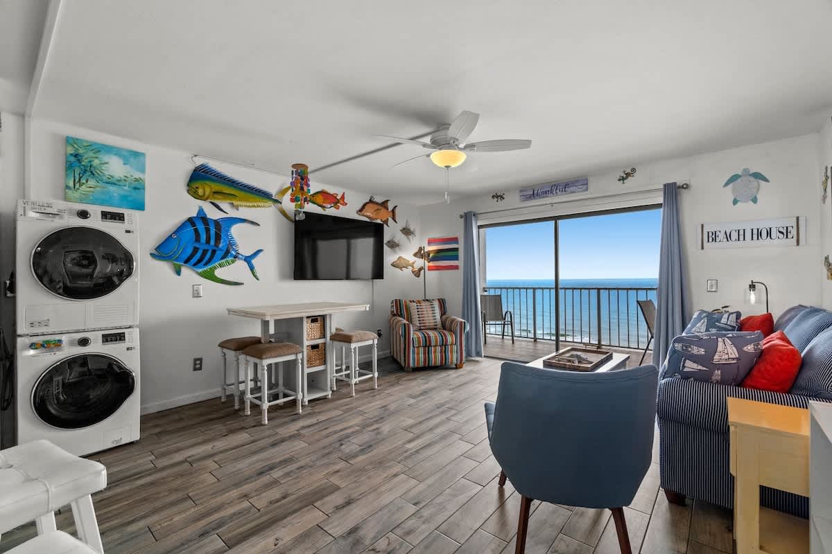 The Summit 1213 – Cozy living area with ocean view, seating, dining table, wall-mounted TV, and washer/dryer units.