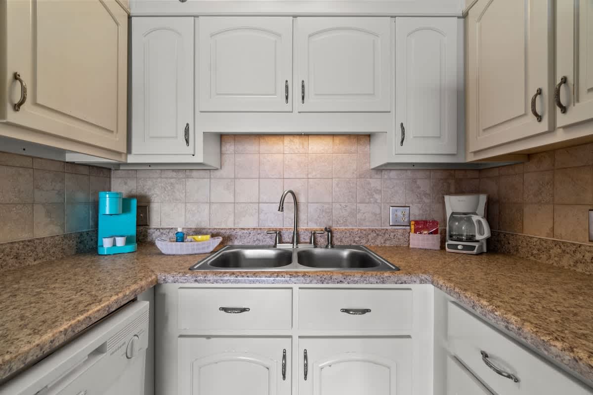 The Summit 1213 – Kitchen with double sink, granite countertops, and white cabinets, featuring a coffee maker and colorful storage items.