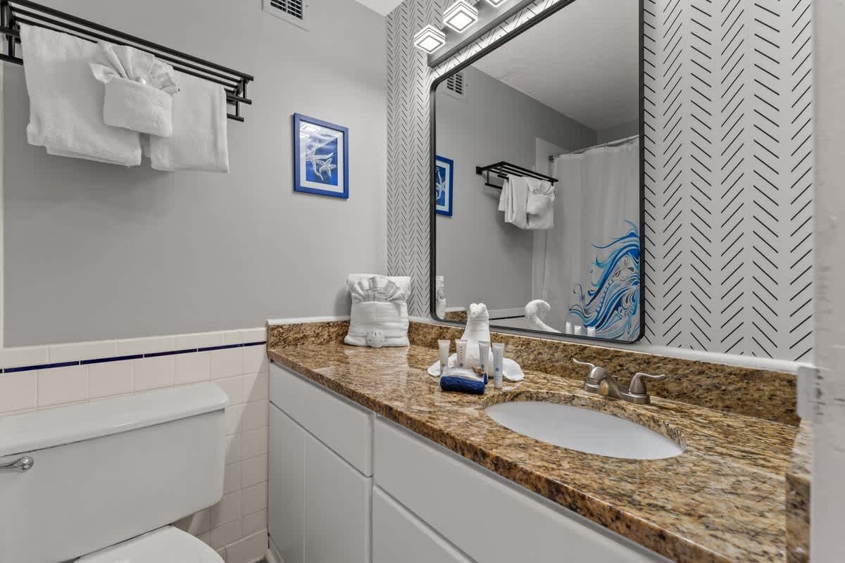 Top Of The Gulf 306 – Bathroom with granite counter, mirror, towels, and decorative shower curtain featuring waves.