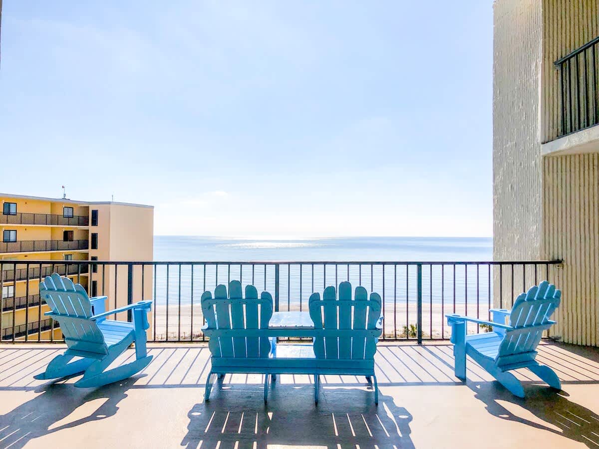 Top Of The Gulf 306 – Balcony with blue Adirondack chairs, ocean view, and nearby building in the background.