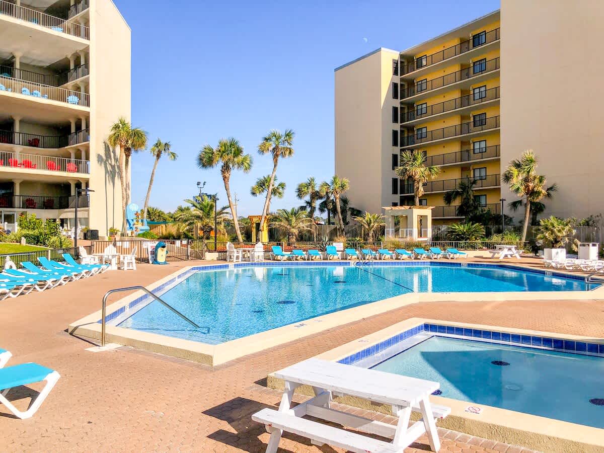 Top Of The Gulf 306 – Outdoor pool area with lounge chairs and palm trees, adjacent to a multi-story building.
