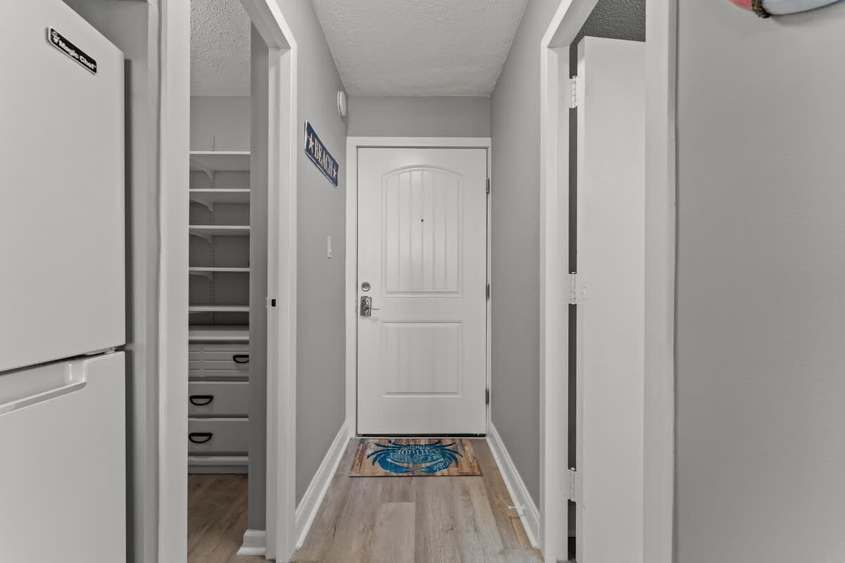 Top Of The Gulf 306 – Hallway entrance with a white door, gray walls, and a refrigerator on the left.