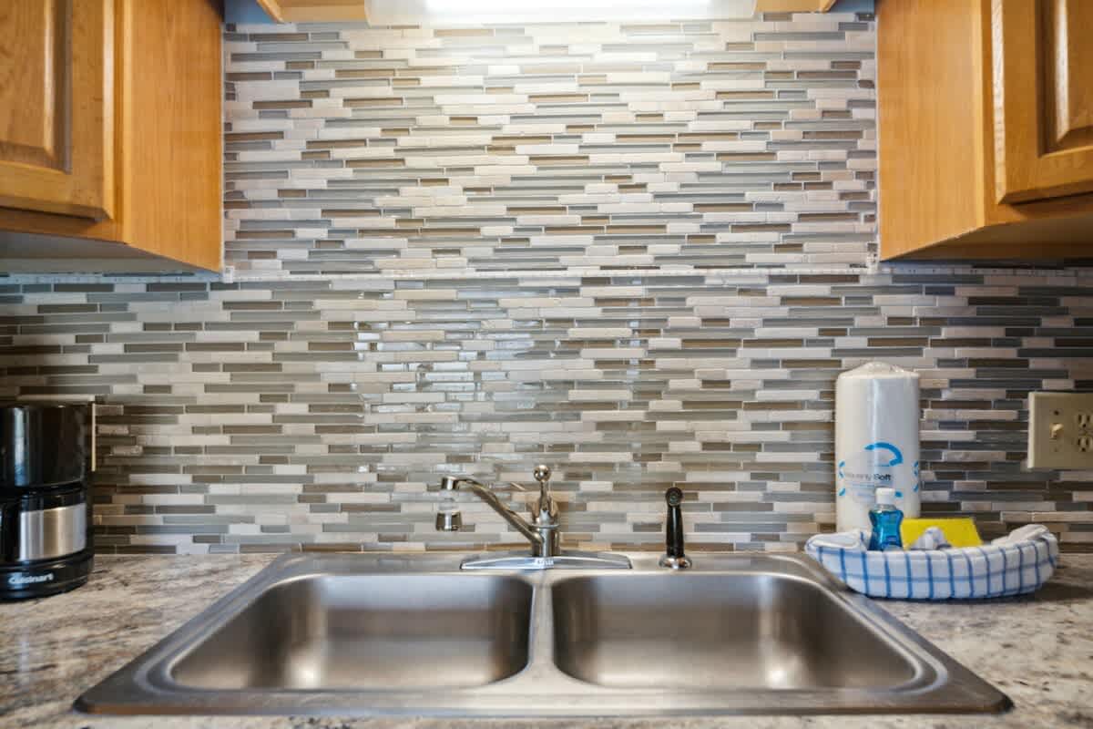 The Summit 817 – Kitchen sink with stainless steel double basin, decorative mosaic backsplash, and countertop items including a coffee maker.