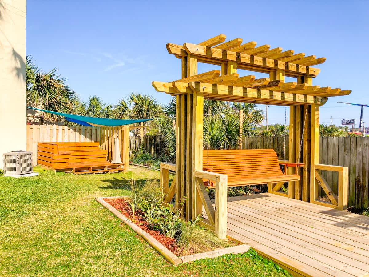 Top Of The Gulf 704 – Outdoor patio with wooden swing, landscaped area, and shaded seating.