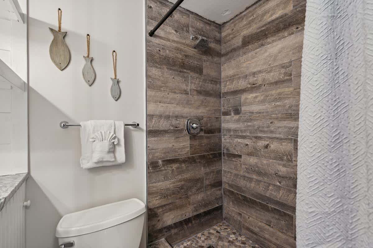 Top Of The Gulf 704 – Bathroom with wooden accent walls, shower, toilet, and decorative fish wall hangings.