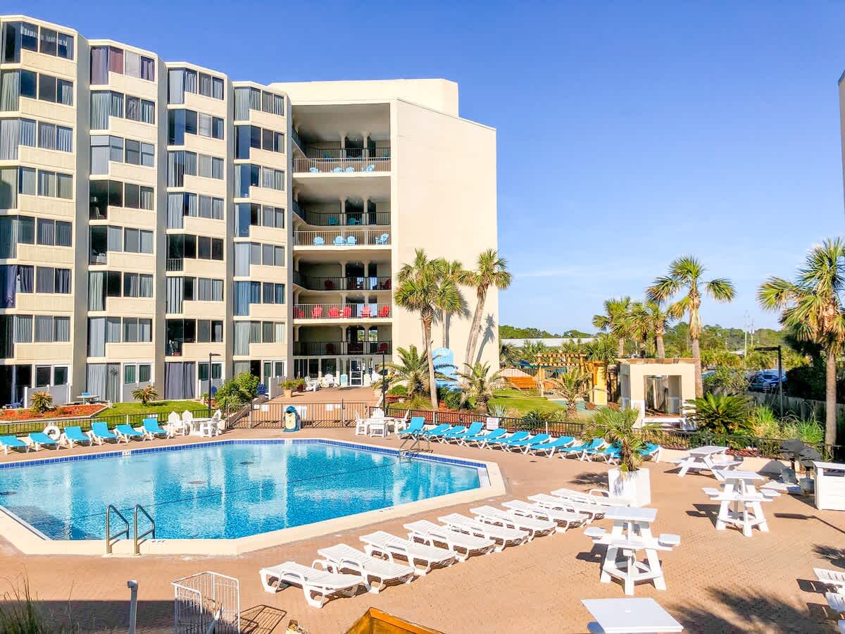 Top Of The Gulf 704 – Pool area with lounge chairs, palm trees, and a building featuring balconies overlooking the pool.