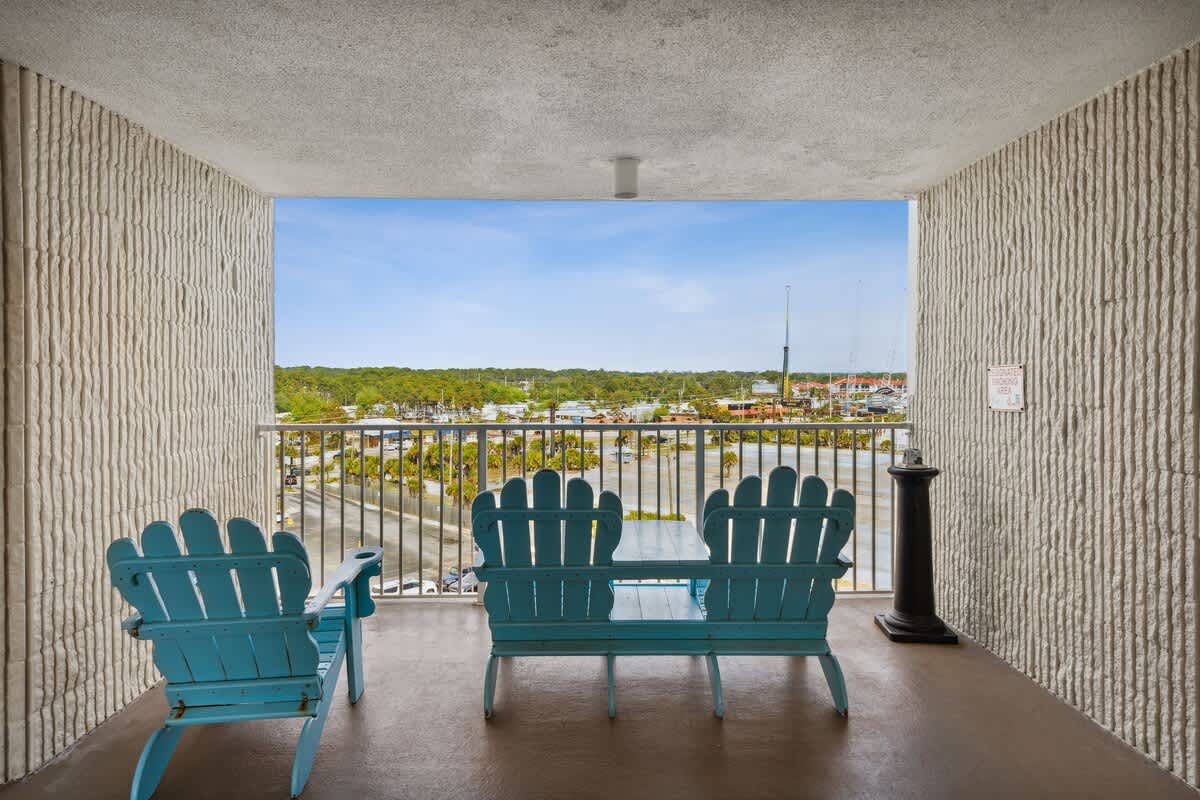 Top Of The Gulf 704 – View from balcony with two turquoise chairs overlooking a marina and tree line.