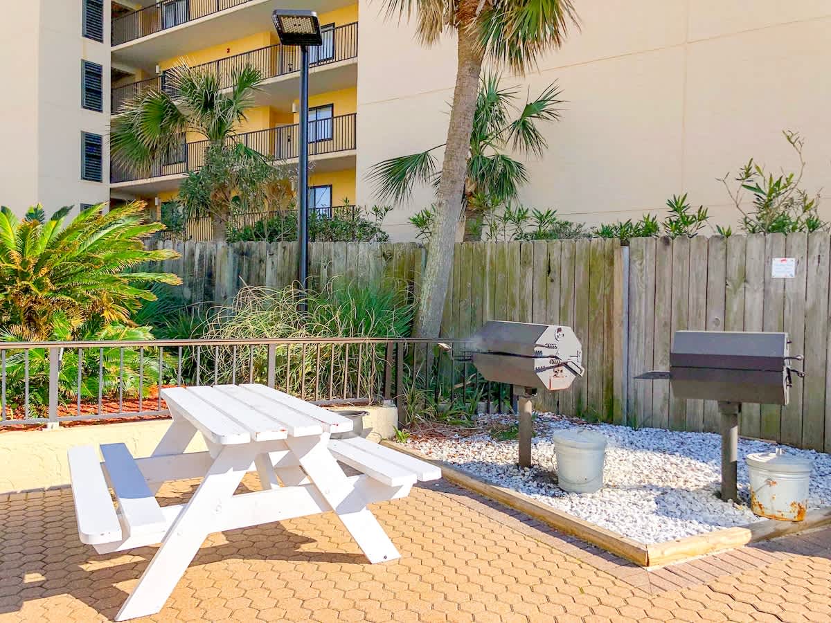 Top Of The Gulf 704 – Picnic table with grilling area and palm trees, near a multi-story building.