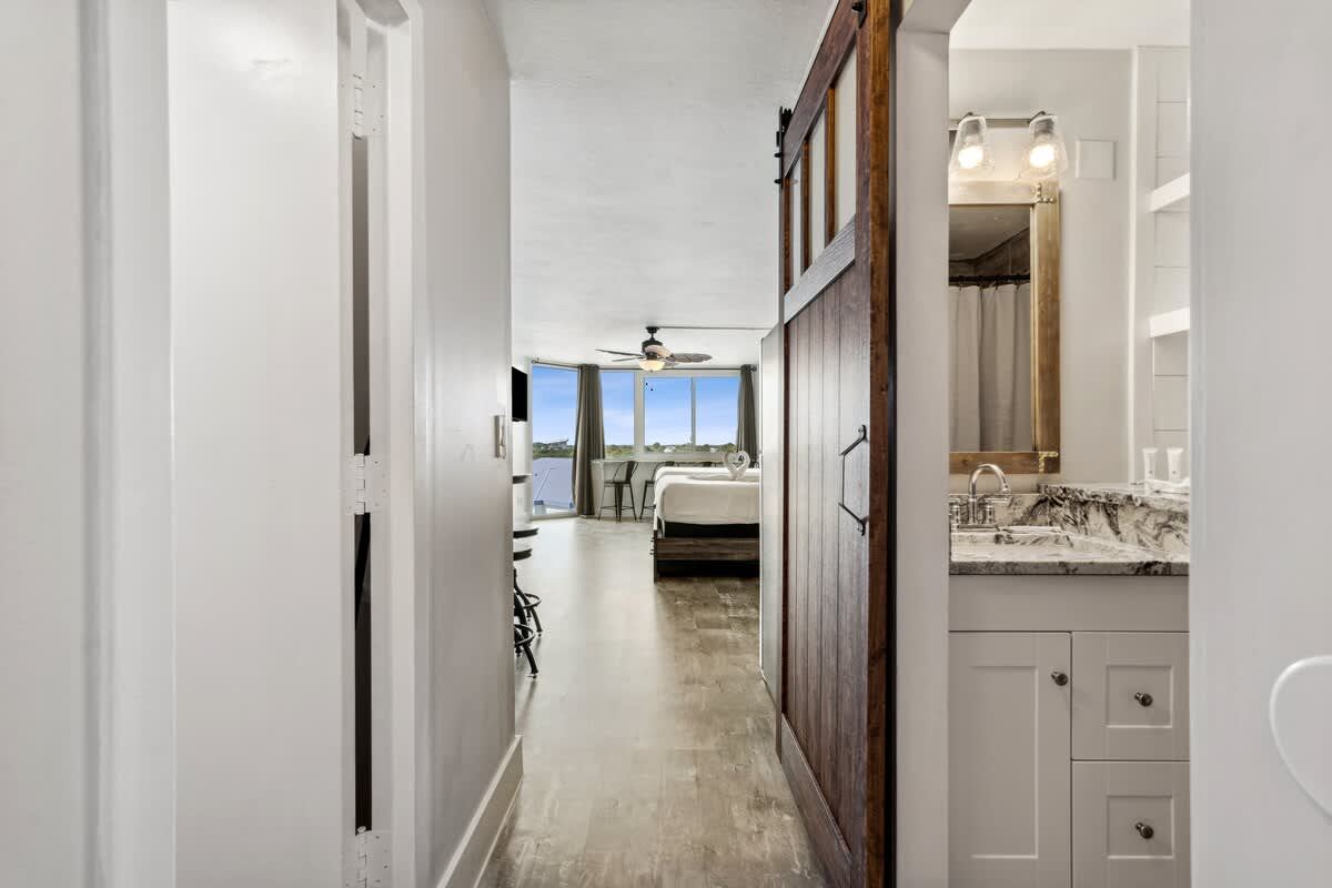 Top Of The Gulf 704 – Wide view of a rental unit showing a hallway, bathroom sink, bedroom area, and large windows with a water view.