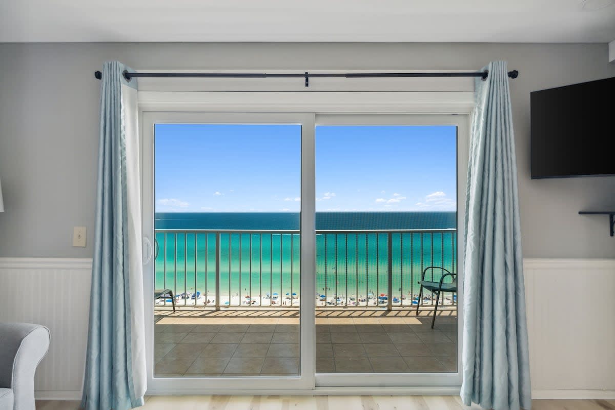 The Summit 1222 – Balcony view of the beach and ocean, with sliding glass doors and light blue curtains.