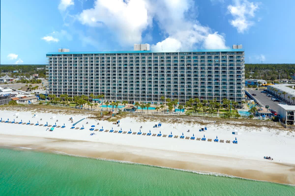 The Summit 725 – Panoramic view of a large beachfront hotel with pool and lounge chairs on white sand.