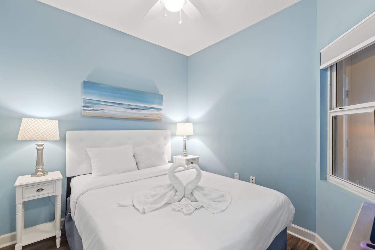 Seychelles 205 – Cozy bedroom with queen bed, blue walls, nightstands, lamps, and a window with an ocean view.