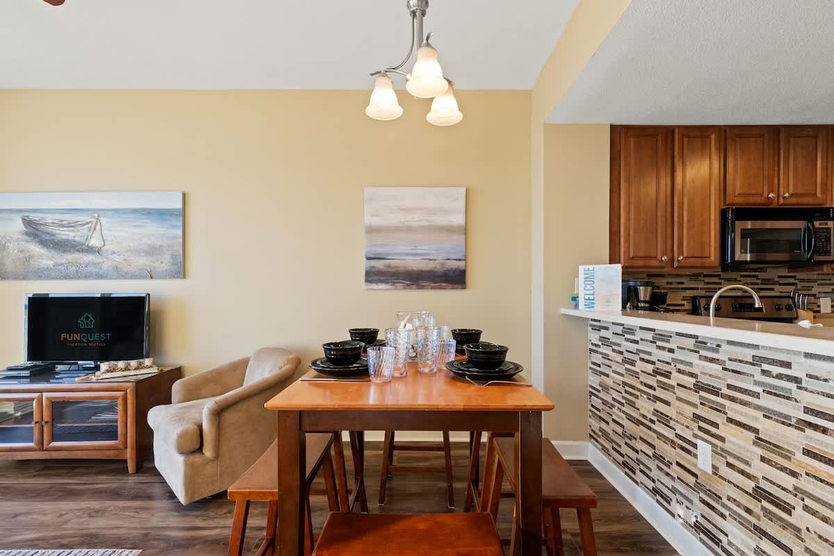 Seychelles 205 – Bright dining area with a wooden table, black dishware, and a connected kitchen featuring modern appliances.