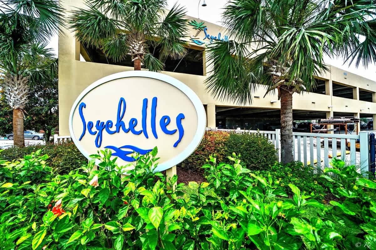 Seychelles 205 – Sign with "Seychelles" name surrounded by greenery and palm trees near a parking structure.