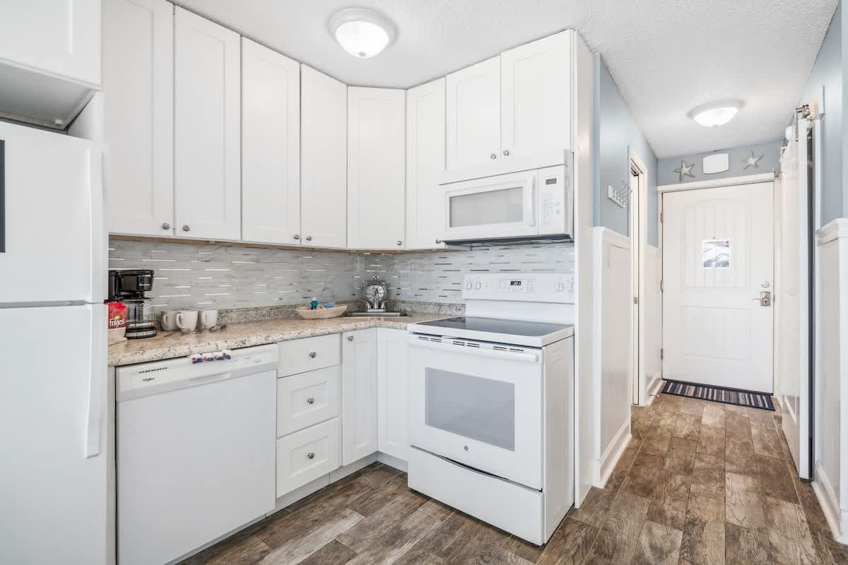 Top Of The Gulf 613 – Compact kitchen featuring white cabinetry, stainless appliances, and granite countertops with a door leading to the hallway.