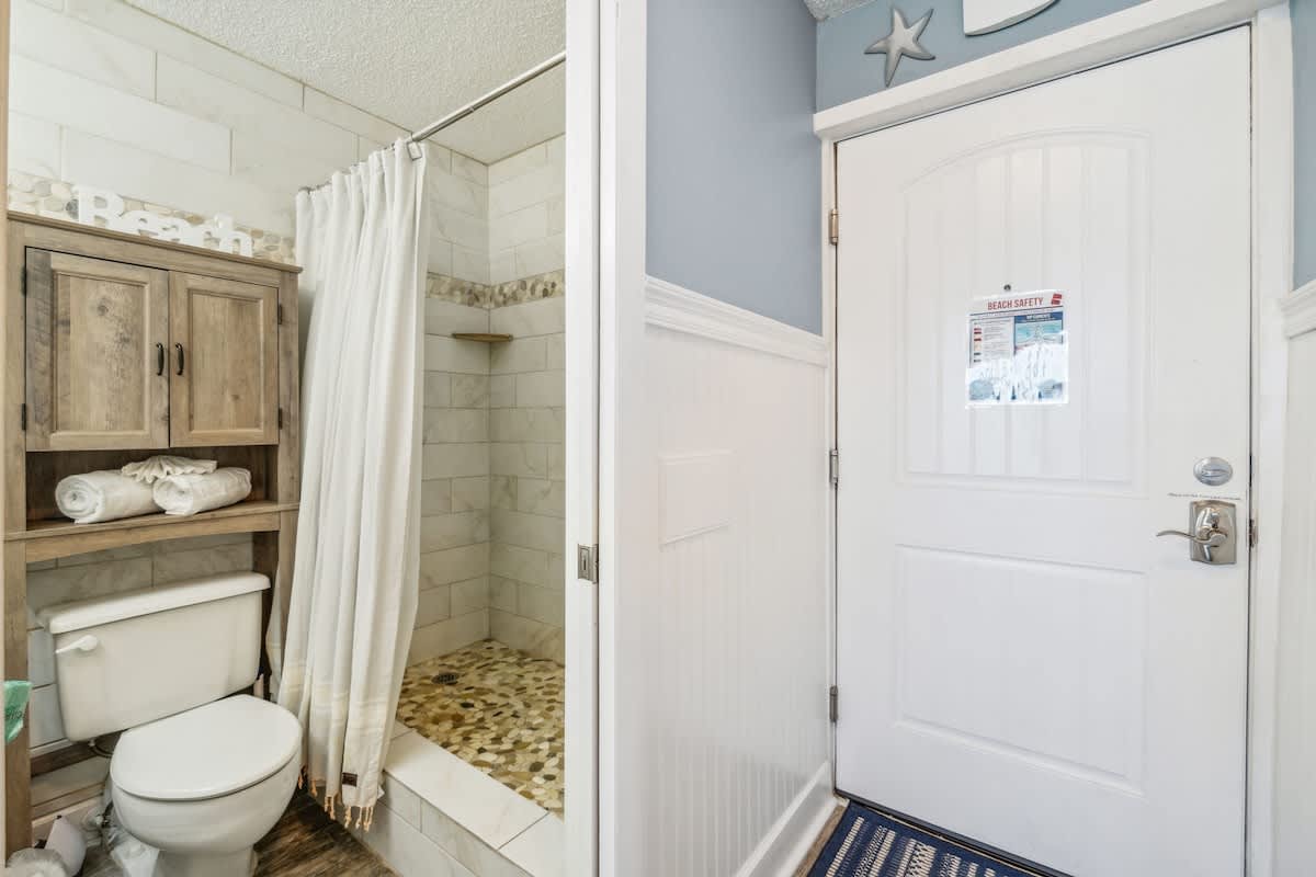 Top Of The Gulf 613 – Bathroom with shower, toilet, shelves, and blue wall; door with safety notice visible.