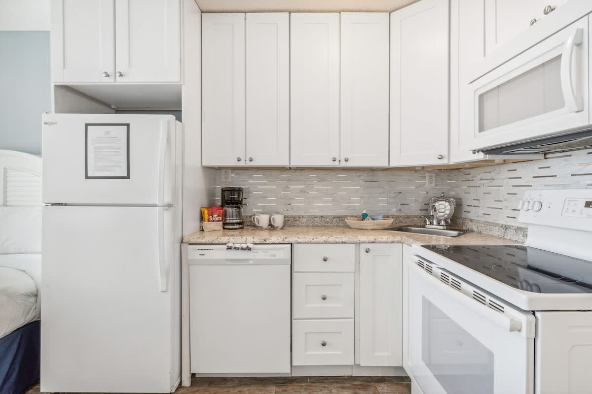 Top Of The Gulf 613 – Compact kitchen with white cabinets, modern appliances, and a view of a bed.